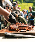 Person cutting barbecue ribs on a wooden board with a crowd in the background