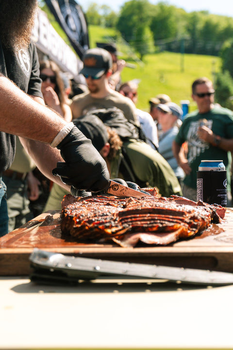 Person cutting barbecue ribs on a wooden board with a crowd in the background