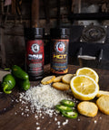 Two bottles of hot sauce on a wooden surface with jalapeños, lemon slices, and crackers.