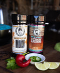 Two bottles of seasoning on a wooden surface with vegetables.