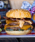 Double cheeseburger with bacon and pickles on a sesame seed bun, with a checkered cloth in the background.