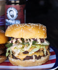 Deluxe burger with multiple layers of meat, cheese, and vegetables on a sesame seed bun, with a coffee cup in the background.