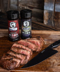 Sliced steak on a wooden cutting board with two spice containers and a knife.