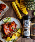 Plated chicken dish with grilled pineapple, lime, and a bottle of barbecue sauce on a wooden surface.