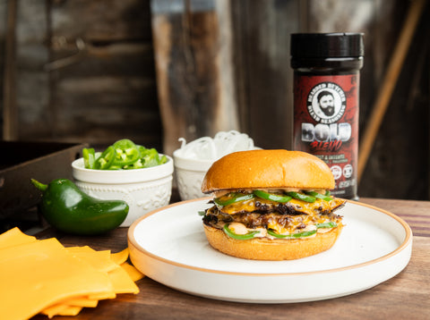 Burger on a plate with jalapeños, cheese, and a shaker of Bearded Butcher Bold Blend on a wooden table.