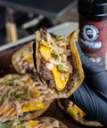 Hand holding a loaded taco with cheese and sauce, with a seasoning in the background.