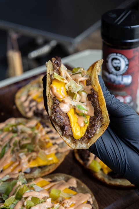Hand holding a loaded taco with cheese and sauce, with a seasoning in the background.